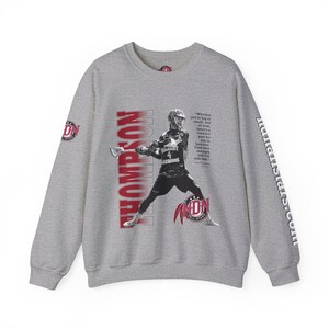 May include: Gray sweatshirt with a lacrosse player graphic and the text "Thompson" printed vertically down the left side. The player is wearing a red and white jersey with the number 4 on it. The text "Whether you're big or small, fast or slow there's a creative part for lacrosse. Find your strength and roll with that." is printed on the right side of the sweatshirt. The text "NON All Stars" is printed on the bottom right of the sweatshirt. The text "nallstars.coLLL" is printed vertically down the right sleeve.