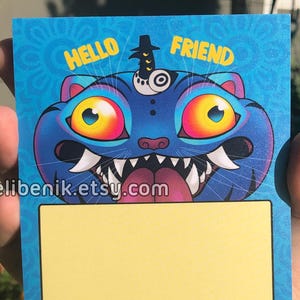 以下が含まれることがあります： 青いノートパッド。漫画の猫のイラストが描かれています。猫は大きく黄色と赤の目、鋭い歯、小さな帽子をしています。「HELLO FRIEND」の文字が猫の上にあります。