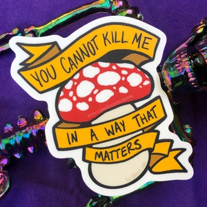 Könnte beinhalten: Ein weißer Aufkleber mit einem gelben Banner, auf dem "You cannot kill me in a way that matters" steht. Das Banner ist um einen rot-weißen Pilz mit weißen Punkten gewickelt. Der Aufkleber befindet sich auf einem violetten Hintergrund.