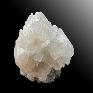 May include: A close-up of a white, crystalline mineral formation. The mineral has a rough, textured surface with many small, pointed crystal structures. The base of the mineral is a dark gray color, and the background is a gradient of black to gray.