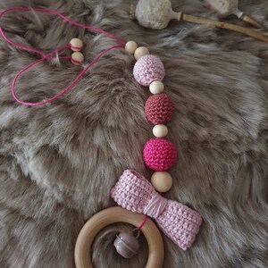 Könnte beinhalten: Ein rosa-brauner gehäkelter Babyschnuller mit Holzperlen und einem Holzring mit einer Glocke. Der Schnuller liegt auf einer flauschigen braunen Oberfläche.