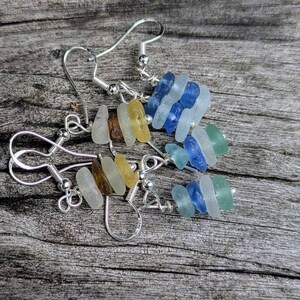 May include: Handmade earrings with sea glass in blue, green, yellow, and brown hues. The sea glass pieces are strung on silver-coloured hooks and wire, creating a unique look. The earrings are displayed on a wooden surface.