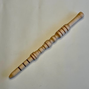 Puede incluir: Una varita de madera de color claro con un mango redondeado y un extremo cónico. La varita presenta una serie de anillos tallados y un diseño texturizado y acanalado a lo largo de su longitud. La varita mide aproximadamente 25 cm de largo.