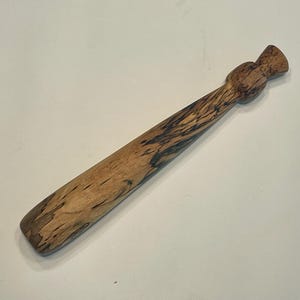 Puede incluir: Un calzador de madera con una veta natural de color marrón claro y vetas oscuras e irregulares. El extremo del mango tiene forma de pomo redondeado, mientras que el extremo opuesto es plano y ligeramente curvado para su uso.