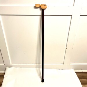 May include: A walking cane featuring a dark brown, slender cylindrical shaft and a light-colored, smooth wooden handle shaped like a stylized bird head. The base of the cane is finished with a black rubber tip for stability and grip.