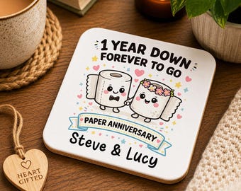 May include: A square coaster with rounded corners featuring a cartoon illustration of two toilet paper rolls holding hands. The text reads 1 YEAR DOWN FOREVER TO GO, PAPER ANNIVERSARY, and Steve & Lucy, surrounded by small colourful hearts and stars.