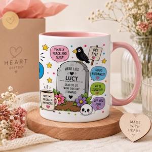 May include: A white ceramic mug with a pink interior and handle, featuring a tombstone graphic inscribed with "HERE LIES LUCY" and "DEAD TO US FROM THIS DAY FORWARD." The design includes a black bird, a skull, colourful flowers, and speech bubbles with text.