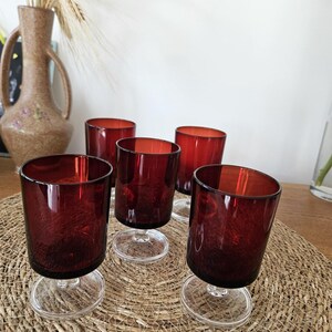 Peut inclure: Un ensemble de cinq verres à boire assortis avec des bols translucides rouge foncé et des pieds en verre transparent avec des bases circulaires. Les verres présentent une forme cylindrique avec une finition lisse, posés sur un tapis tissé.