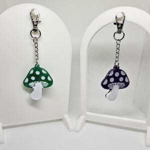 May include: Two mushroom keychain charms, one green with white dots and one purple with white dots. Both charms are attached to silver chains and key rings.