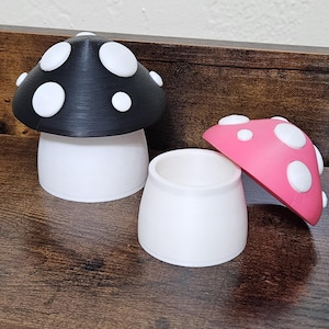 May include: A whimsical mushroom-shaped container with a black cap and white spots, a white base, and a separate pink cap with white spots. The container is sitting on a wooden surface. The mushroom-themed decor is a unique addition to any room.