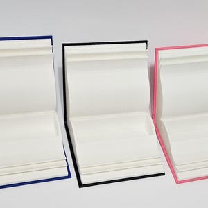 May include: Three open book-shaped containers with white pages and colored edges. The containers have blue, black, and pink edges. The containers are on a white surface.