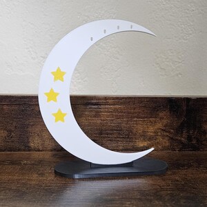 May include: A white crescent moon-shaped jewelry holder with three yellow stars. The moon has small holes for hanging earrings and sits on a black oval base. The jewelry holder is displayed on a dark wooden surface.