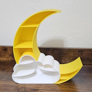 May include: A yellow crescent moon-shaped shelf with three interior shelves. A white cloud-shaped shelf is attached to the bottom of the moon. The shelf is made of wood and sits on a wooden surface.