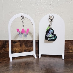 May include: Two white arched display stands. One holds a pink and silver glitter letter "W" keychain. The other holds a black heart-shaped keychain with a butterfly design and a green tassel. Both keychains are attached with silver chains.
