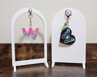 May include: Two white arched display stands. One holds a pink and silver glitter letter "W" keychain. The other holds a black heart-shaped keychain with a butterfly design and a green tassel. Both keychains are attached with silver chains.