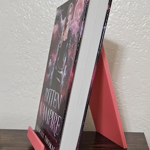 May include: A book titled "Bitten Vampire" by Rogan Thomas, displayed on a coral-colored book stand. The book cover features a woman with a dark background and the title in white. The stand is made of a solid material.