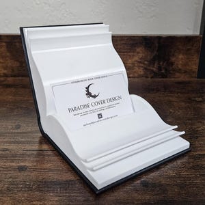 May include: White book cover design business card holder with a black base. The business card reads "Paradise Cover Design" and includes the text "Specializing in unique fantasy, pop and fantasy themed covers, paranormal, fairy tale retellings, and dark fantasy." The business card also includes the email address "melany@paradisecoverdesign.com".
