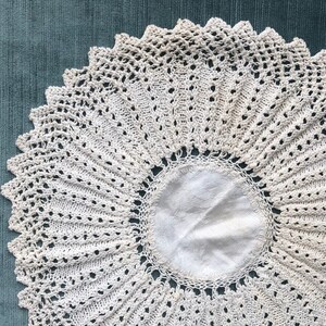 Puede incluir: Un tapete circular de color crema con un diseño de encaje de ganchillo. El tapete presenta un círculo central sólido rodeado por un intrincado patrón calado. Los bordes están festoneados, lo que añade un toque decorativo. El tapete está colocado sobre una superficie de color verde azulado.