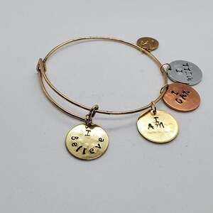 May include: A gold-toned wire bangle bracelet featuring four round metal charms. The charms are stamped with the phrases I BELIEVE, I AM, I CAN, and I WILL. The charms are finished in gold, copper, and silver tones, hanging from the circular band.