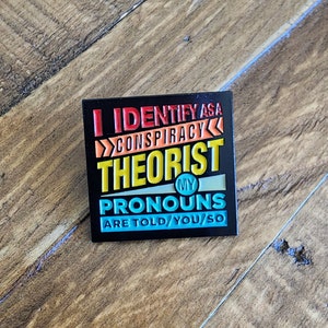 May include: Black enamel pin with the text "I identify as a conspiracy theorist. My pronouns are told/you/so" in colorful letters.