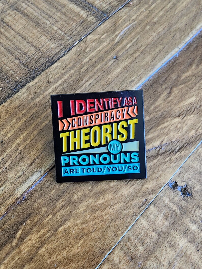 May include: Black enamel pin with the text "I identify as a conspiracy theorist. My pronouns are told/you/so" in colorful letters.