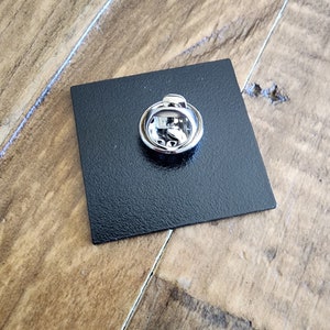 May include: The back of a black square pin with a silver pin back.