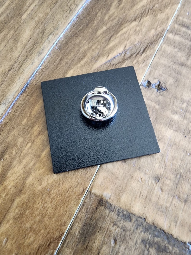 May include: The back of a black square pin with a silver pin back.