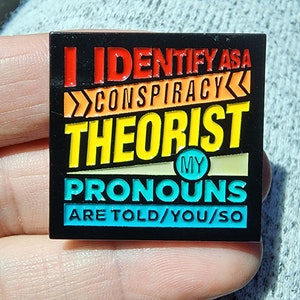 May include: A black enamel pin with a colorful design. The pin says "I identify as a conspiracy theorist. My pronouns are told/you/so."