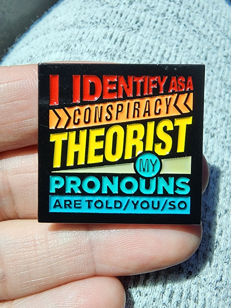 May include: A black enamel pin with a colorful design. The pin says "I identify as a conspiracy theorist. My pronouns are told/you/so."