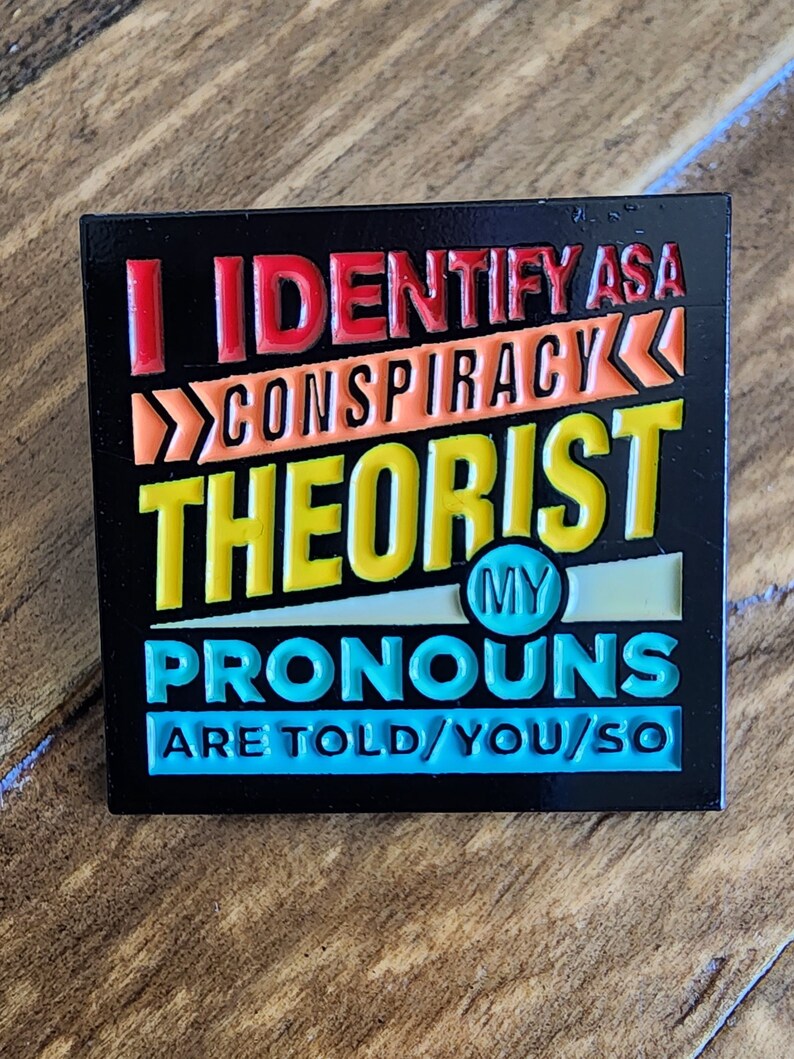 May include: Black enamel pin with the text "I identify as a conspiracy theorist. My pronouns are told/you/so" in colorful letters.