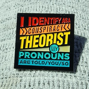 May include: Black enamel pin with a rainbow color scheme. The pin says "I identify as a conspiracy theorist. My pronouns are told/you/so."