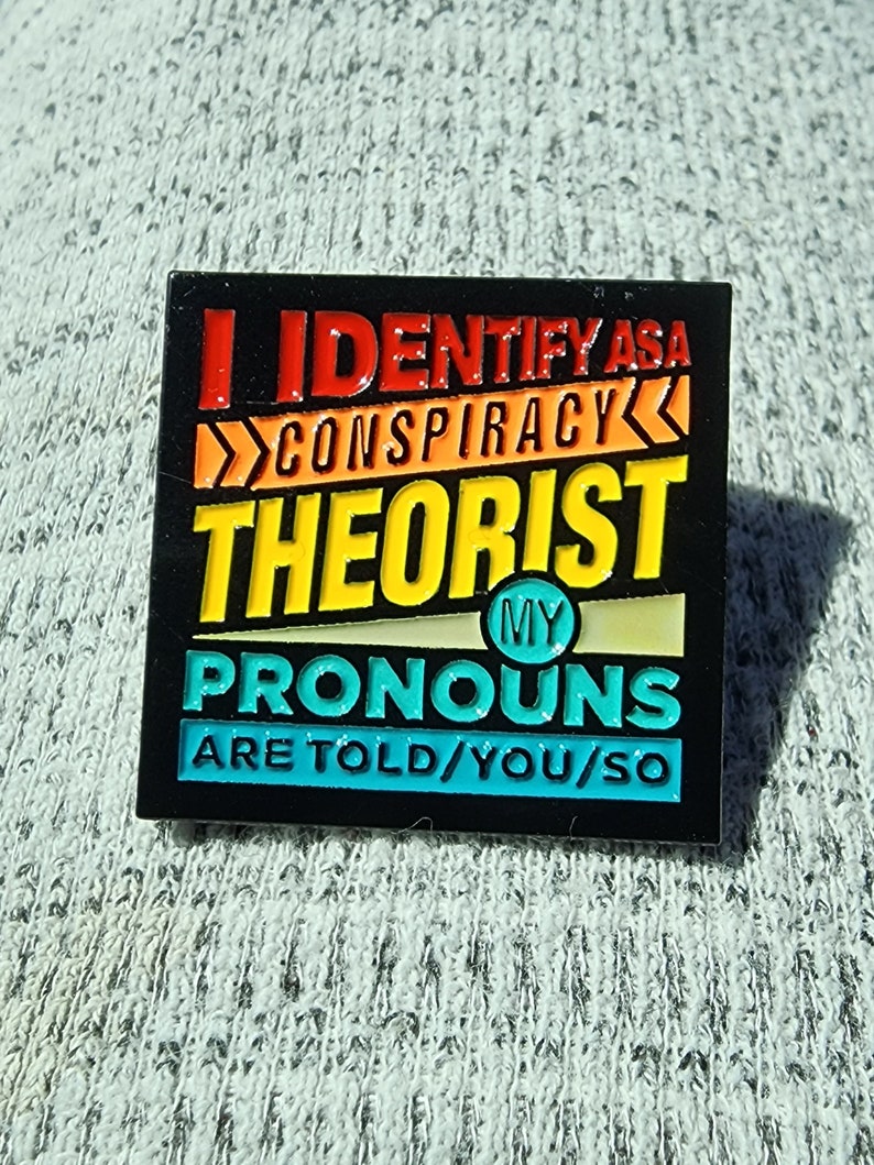 May include: Black enamel pin with a rainbow color scheme. The pin says "I identify as a conspiracy theorist. My pronouns are told/you/so."