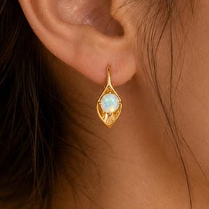 May include: A gold leverback earring featuring a central round iridescent opal stone set within a smooth, petal-shaped metal frame that tapers to a point at the bottom, creating a delicate and polished appearance on the earlobe.