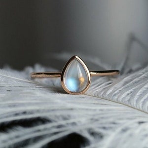 May include: A gold ring featuring a pear-shaped moonstone set in a smooth bezel. The gemstone displays a soft, iridescent blue glow, and the band is thin and polished. The ring is positioned on a white, textured feather-like surface.