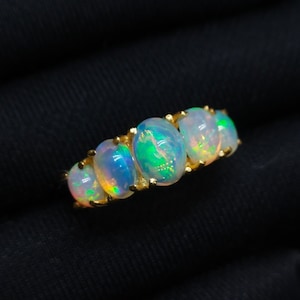 Könnte beinhalten: Ein Goldring mit fünf ovalen Cabochon-Opalen, die in einer Reihe angeordnet sind. Jeder Opal zeigt eine durchscheinende weiße Basis mit lebhaften grünen, orangen und blauen Farbspielen, befestigt durch kleine Goldkrappen am Ringband.