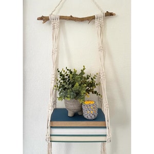 May include: A macrame wall shelf with a natural wood branch, hanging from the ceiling. The shelf is holding a small potted plant, a small gray crocheted candle holder, and a stack of books.