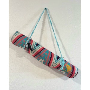 Op de afbeelding: Een kleurrijke yogamat met een blauwe en witte band die aan een witte muur hangt. De mat heeft een geometrisch patroon met roze, blauwe en witte kleuren. De band is gemaakt van macramé en heeft een sleutel eraan.