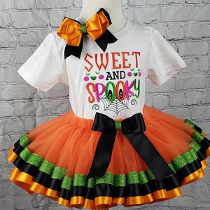 K&ouml;nnte beinhalten: Ein orangefarbener und schwarzer T&uuml;ll-Tutu-Rock mit schwarzem und gr&uuml;nem Bandbesatz. Ein wei&szlig;es T-Shirt mit dem Text "Sweet and Spooky" in rosa, gr&uuml;n und orangefarbenen Buchstaben. Eine gro&szlig;e orangefarbene und schwarze Schleife befindet sich oben auf dem Shirt.
