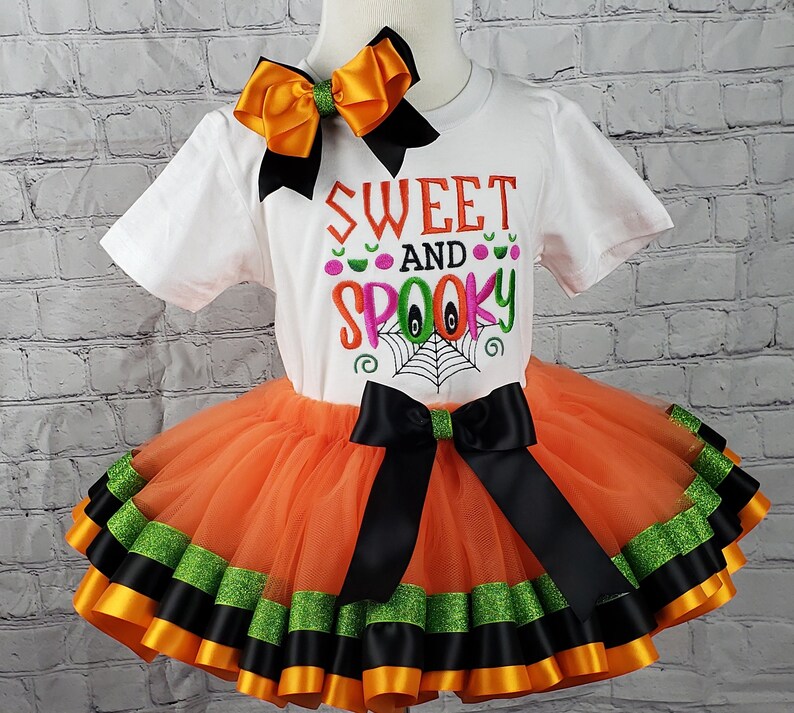 K&ouml;nnte beinhalten: Ein orangefarbener und schwarzer T&uuml;ll-Tutu-Rock mit schwarzem und gr&uuml;nem Bandbesatz. Ein wei&szlig;es T-Shirt mit dem Text "Sweet and Spooky" in rosa, gr&uuml;n und orangefarbenen Buchstaben. Eine gro&szlig;e orangefarbene und schwarze Schleife befindet sich oben auf dem Shirt.