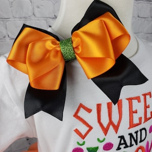 K&ouml;nnte beinhalten: Eine gro&szlig;e orangefarbene und schwarze Schleife mit gr&uuml;nem Glitzer in der Mitte. Die Schleife befindet sich auf einem wei&szlig;en Shirt mit dem aufgen&auml;hten Text "Sweet and".