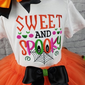 K&ouml;nnte beinhalten: Ein wei&szlig;es T-Shirt mit dem Text "Sweet and Spooky", bestickt in Orange, Pink, Gr&uuml;n und Schwarz. Das Shirt wird mit einem orangefarbenen T&uuml;ll-Tutu und einer schwarzen Schleife mit gr&uuml;nem Glitzer kombiniert.