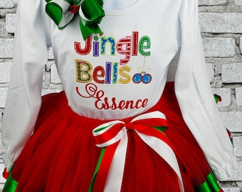 May include: A white long-sleeved shirt with the text "Jingle Bells" in colorful letters and the name "Essence" underneath. The shirt is paired with a red tulle skirt with green and white ribbon trim.