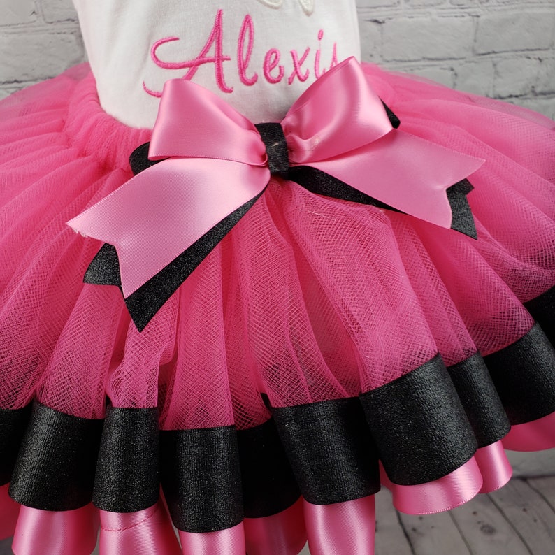 May include: Pink tulle tutu skirt with black satin ribbon trim and a large pink satin bow. The skirt has the name "Alexis" embroidered on the front.
