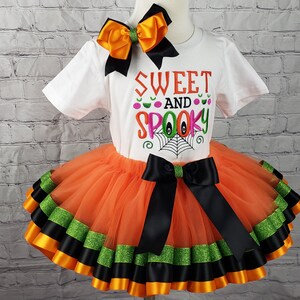 K&ouml;nnte beinhalten: Ein orangefarbener und schwarzer T&uuml;ll-Tutu-Rock mit schwarzem und gr&uuml;nem Bandbesatz. Ein wei&szlig;es T-Shirt mit dem Text "Sweet and Spooky" in rosa, gr&uuml;n und schwarzer Stickerei. Eine schwarze und orange Schleife befindet sich oben auf dem Shirt.