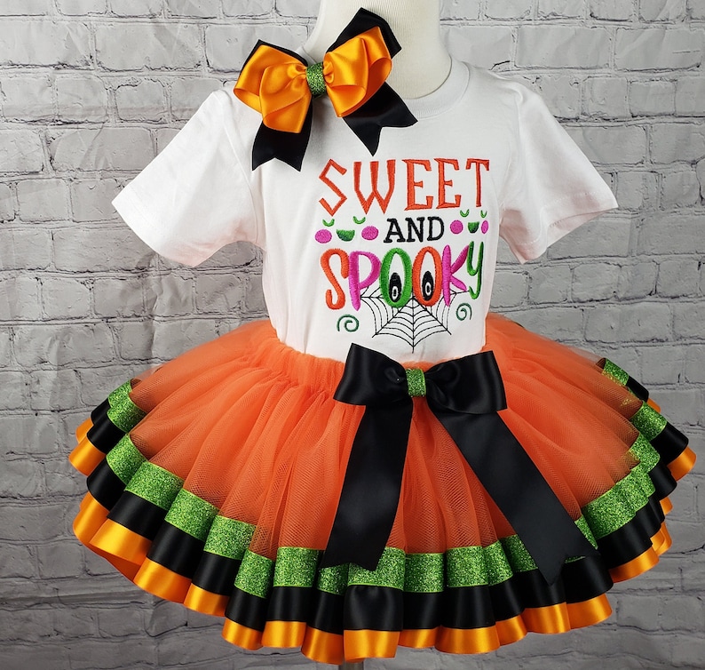 K&ouml;nnte beinhalten: Ein orangefarbener und schwarzer T&uuml;ll-Tutu-Rock mit schwarzem und gr&uuml;nem Bandbesatz. Ein wei&szlig;es T-Shirt mit dem Text "Sweet and Spooky" in rosa, gr&uuml;n und schwarzer Stickerei. Eine schwarze und orange Schleife befindet sich oben auf dem Shirt.