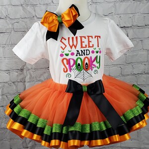 K&ouml;nnte beinhalten: Ein orangefarbener und schwarzer T&uuml;ll-Tutu-Rock mit schwarzem und gr&uuml;nem Bandbesatz. Ein wei&szlig;es T-Shirt mit dem Text "Sweet and Spooky" in Pink, Gr&uuml;n und Orange. Eine gro&szlig;e orange Schleife befindet sich auf dem Shirt.