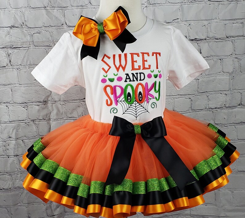 K&ouml;nnte beinhalten: Ein orangefarbener und schwarzer T&uuml;ll-Tutu-Rock mit schwarzem und gr&uuml;nem Bandbesatz. Ein wei&szlig;es T-Shirt mit dem Text "Sweet and Spooky" in Pink, Gr&uuml;n und Orange. Eine gro&szlig;e orange Schleife befindet sich auf dem Shirt.