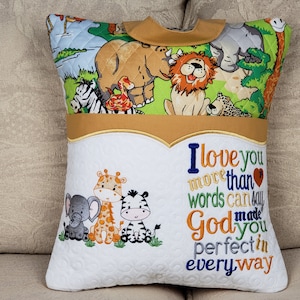 May include: A white pillow with a brown and green quilted fabric on the front. The front features a variety of animal illustrations, including a zebra, rhino, giraffe, and lion. The back of the pillow is white with a gold trim and the text "I love you more than words can say God made you perfect in every way".