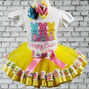 May include: Yellow tulle tutu skirt with multiple layers of yellow and white ribbon with an Easter egg design. The skirt is paired with a white t-shirt featuring three pastel-colored bunny rabbits holding Easter eggs and the text "Happy Easter".