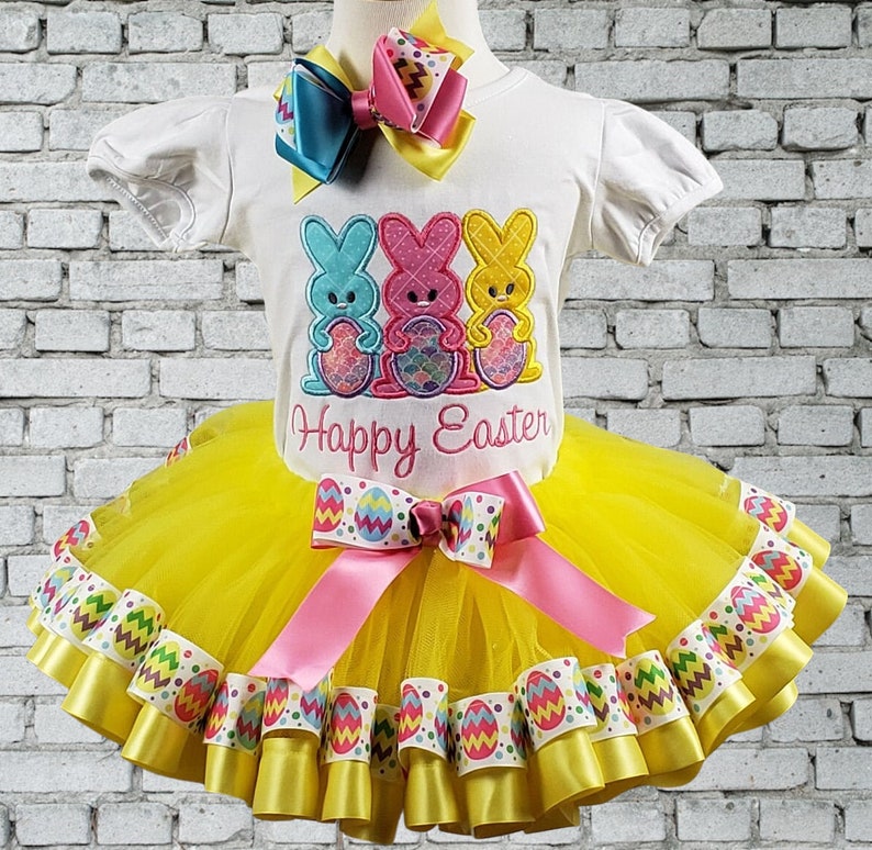 May include: Yellow tulle tutu skirt with multiple layers of yellow and white ribbon with an Easter egg design. The skirt is paired with a white t-shirt featuring three pastel-colored bunny rabbits holding Easter eggs and the text "Happy Easter".